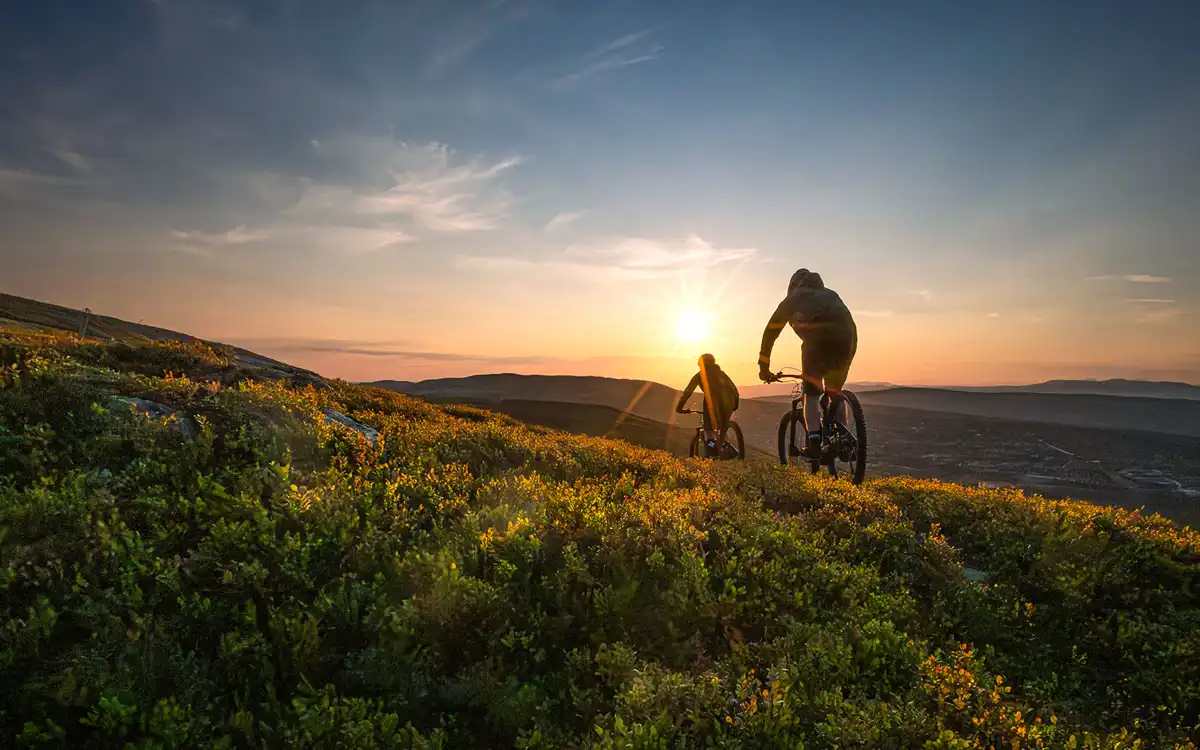 To syklister på terrengsykler sykler over en lyngkledd fjellrygg mot en lav sol, med fjell i bakgrunnen